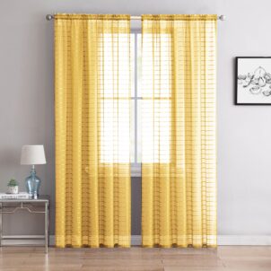 A window adorned with yellow lace curtains, allowing light to filter through. A small lamp and books sit on a side table to the left, and a framed black and white floral artwork hangs on the right. The room features light gray walls and wooden flooring