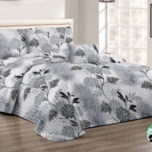 A neatly made bed with a grey floral-patterned quilt and matching pillows, set in a bright room with light walls. A small bedside table with a lamp is on the left. A tall window allows natural light to fill the space.