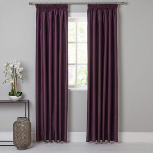 Plain taffeta lined curtains in a rich purple hang on a curtain rod, framing a window with a white frame. Below, a small table with a potted white orchid is positioned against the light gray wall. A large gray vase sits elegantly on the floor beside the table.